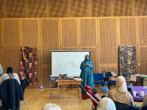 An image showing a meeting held in a hall with wooden floors and woven material wall panels. At the front, a woman wearing a blue hijab is presenting while standing next to a projection screen displaying text that reads 'International Women's Day 2024 or robes. On either side of the room are large artistic displays featuring portraits or collages of diverse faces and images. The scene depicts a community gathering.